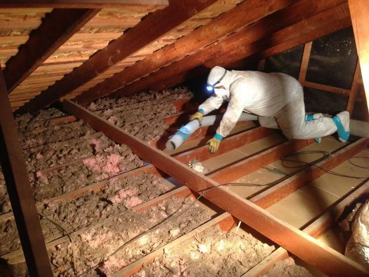 Technician in a protective suit working between attic joists with a flexible duct and old insulation