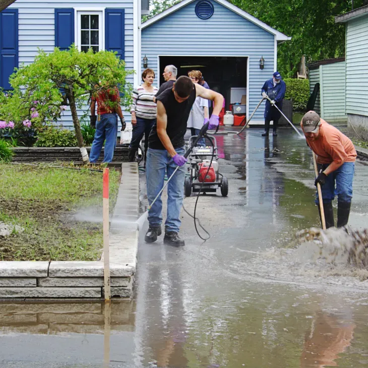 Crew clearing mud and standing flood water from a driveway with hoses and brooms