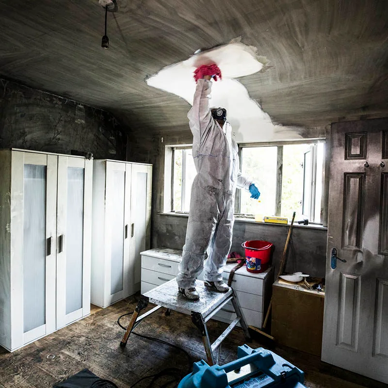 Technician in a protective suit wiping soot off a smoke-blackened bedroom ceiling