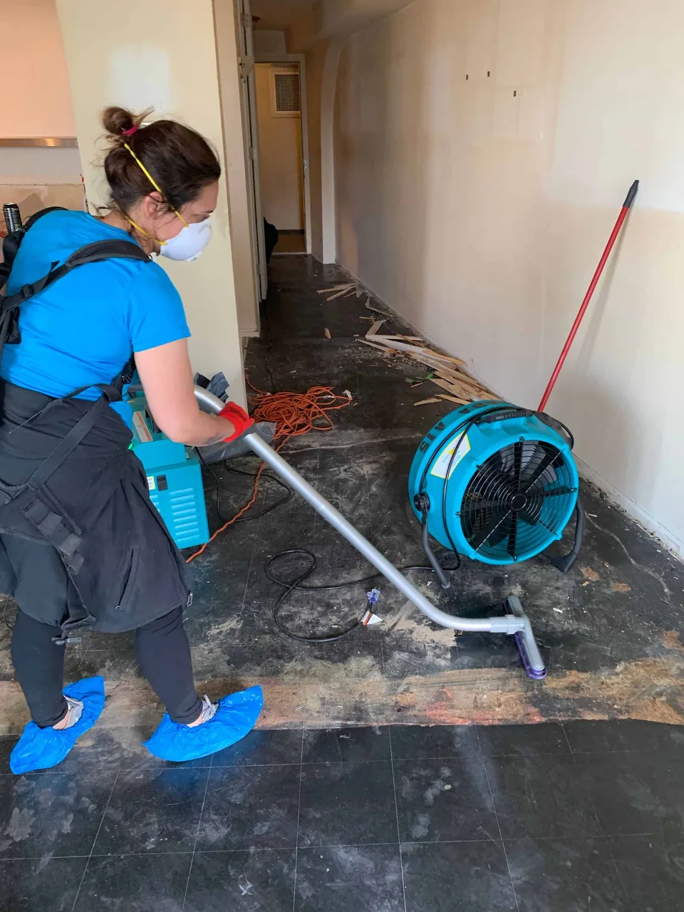 Technician in a mask and shoe covers extracting water from a floor while an air mover dries the hallway