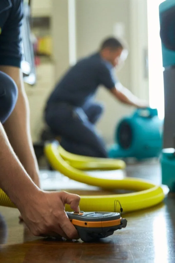 Technician checking a wet hardwood floor with a moisture meter while another sets up an air mover