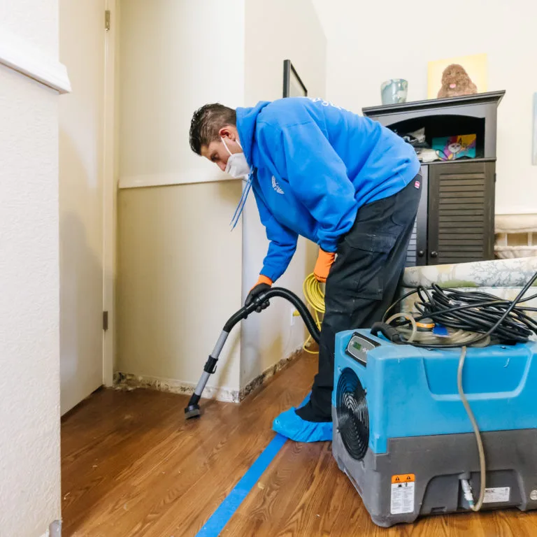 Technician in a mask vacuuming water along a baseboard next to a running dehumidifier