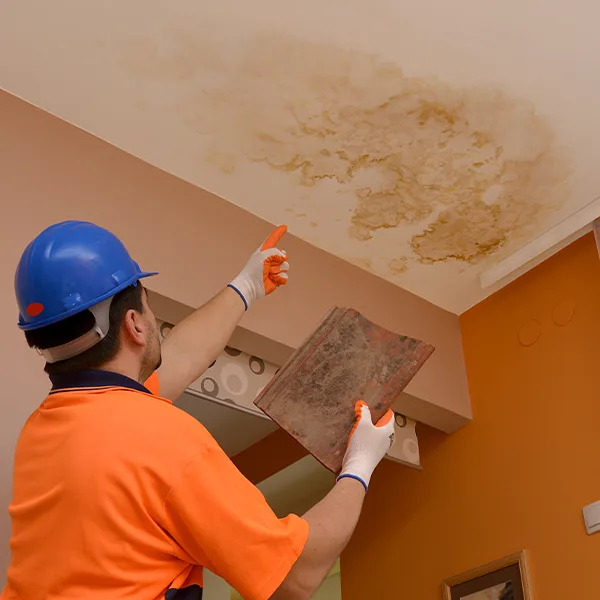 Worker in a hard hat pointing at a water-stained ceiling while holding a damaged roof tile